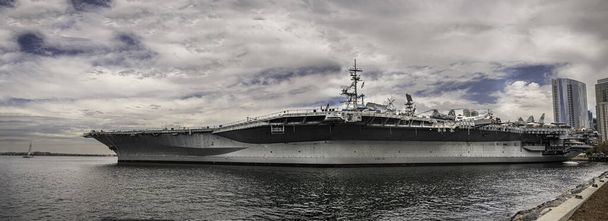 San Diego, Kalifornie, USA - 4. října 2021: USS Midway Museum, Aircraft Carrier. Panoráma lodi kotvila u mola námořnictva pod modrým mrakem a vpředu zátokou vody. Kancelářská budova. - Fotografie, Obrázek