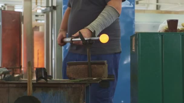 Middle plan glassblowing workshop,stout master,gray T-shirt,blue shorts,long armband,no face,turns yellow hot round blank on hollow tube,supported by metal V-shaped stand fixed on rusty metal box. - Footage, Video