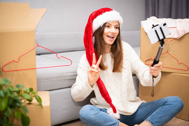 Eine junge schöne Bloggerin mit Weihnachtsmannhut ist in eine neue Wohnung gezogen und nimmt mit einem Handy und einem Selfie-Stick ein Video vor dem Hintergrund von Boxen auf. - Foto, Bild