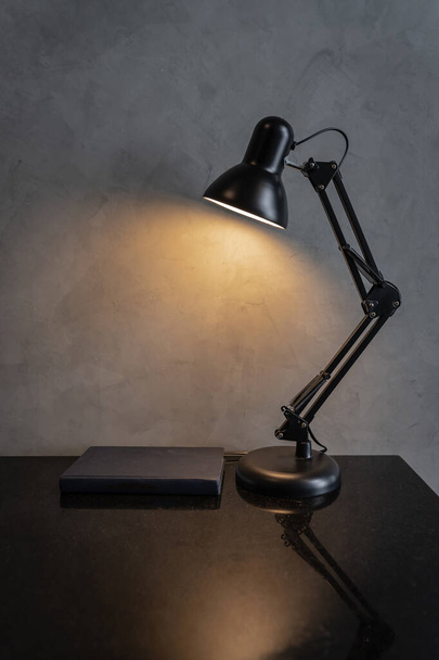 Retro black metal desk lamp or flexible table lamp light bulb on the black table and blue book with cement loft-style wall interior vintage design contemporary. Modern house building concept. - Фото, изображение