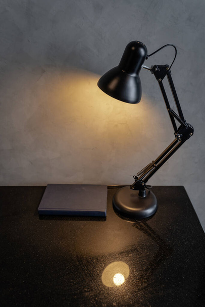 Retro black metal desk lamp or flexible table lamp light bulb on the black table and blue book with cement loft-style wall interior vintage design contemporary. Modern house building concept. - Zdjęcie, obraz