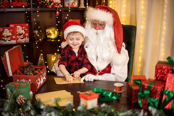 Little child boy visits Santa Claus helps him open envelopes with Christmas wishes. Holidays concept - Foto, imagen