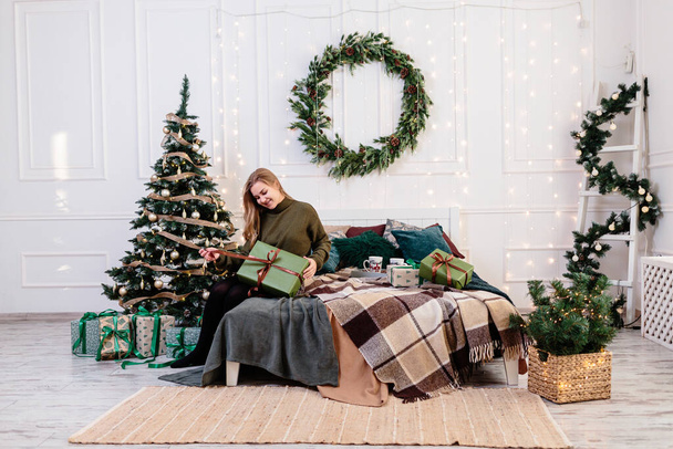 A young woman opens Christmas gifts in a New Year's atmosphere and rejoices. The woman is in a bright, cozy bedroom, beautifully decorated for Christmas. - Photo, Image