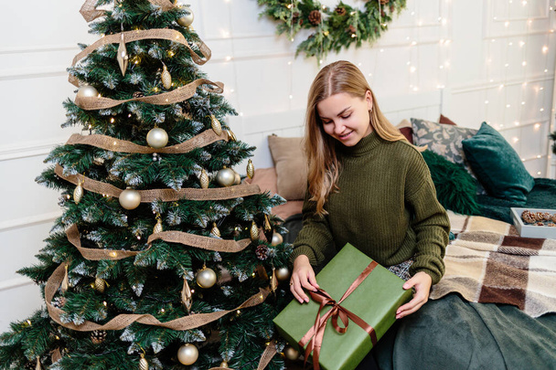 Eine junge Frau öffnet Weihnachtsgeschenke in Neujahrsatmosphäre und freut sich. Die Frau steht in einem hellen, gemütlichen Schlafzimmer, schön weihnachtlich dekoriert. - Foto, Bild