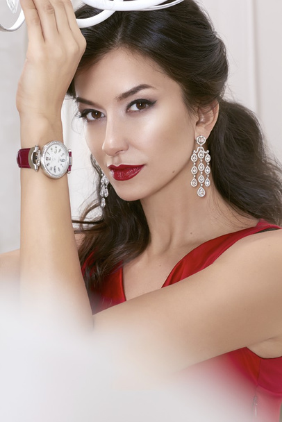 Beautiful sexy woman brunette Brown eyes in a red dress in lavish earrings with diamonds and watches on the Burgundy leather strap evening makeup, red lipstick, raised a hand up - Foto, Imagem