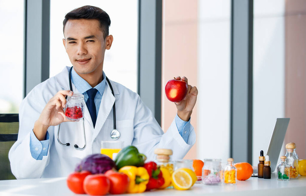Содержание Азиатский мужчина диетолог сидит за столом со свежим яблоком и добавки сравнить их и показать настоящее на камеру - Фото, изображение