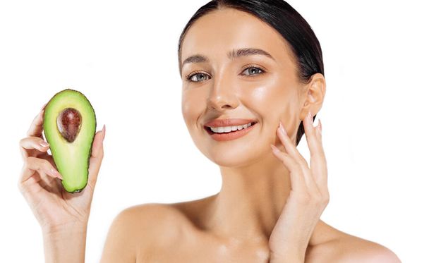 Beautiful happy young woman holding half avocado in front of her face, portrait of attractive woman with perfect makeup on isolated white background, beauty and skin care concept - Foto, Imagem