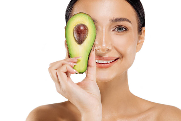 Beautiful woman with clear skin, holding an avocado and covering her eyes, She looks at the camera and smiles softly, her shoulders are bare, on an isolated white background, Beauty and skin care - Fotó, kép