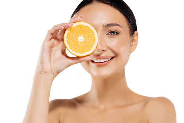 beautiful young woman, with healthy skin, gently touching face, well-groomed complexion, healthy glowing skin, holding half a juicy orange, on an isolated white background, Vitamin C cosmetics concept - Фото, изображение