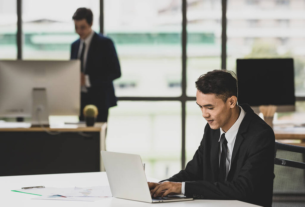 Χαμογελώντας εθνοτική αρσενικό επιχειρηματία δακτυλογράφηση σε netbook και εργάζονται σε έργο στο χώρο εργασίας με θολή αγνώριστη αρσενικό συνάδελφο - Φωτογραφία, εικόνα