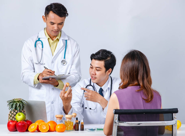 Pozytywne azjatyckie męskie dietetycy z suplementów i zdrowej żywności gromadzenie przy stole z pacjentką na szarym tle - Zdjęcie, obraz