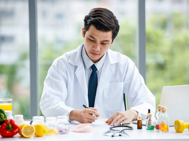 Asiatischer Mann in medizinischer Uniform schaut und denkt über Pillen nach und macht sich Notizen, während er in einer modernen Klinik arbeitet - Foto, Bild