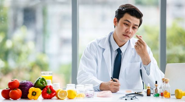 Asiatischer Mann in medizinischer Uniform zeigt Pille und macht sich Notizen bei der Arbeit in moderner Klinik - Foto, Bild