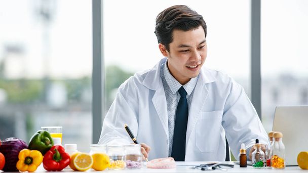 Cheerful Asian medical practitioner smiling and making notes while sitting at table with fruits and vitamins in hospital - Foto, imagen