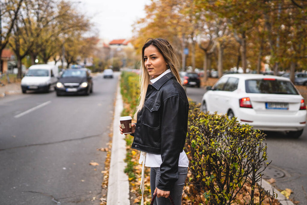 Egy fiatal, komoly kaukázusi nő áll odakint, miközben próbál átkelni az úton ősszel egy csésze kávéval a kezében, valós emberek utánozzák a helyet. - Fotó, kép