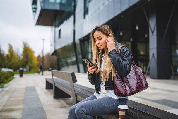 одна молодая кавказка, студентка, сидящая перед университетом осенью в ожидании, пока держит мобильный телефон, читая сообщения или просматривая интернет, реальные люди копируют пространство - Фото, изображение