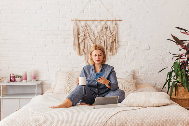 Pijamalı, yatakta dizüstü bilgisayarla çalışan, telefonda konuşan güzel bir genç kadın. İnternetten alışveriş - Fotoğraf, Görsel