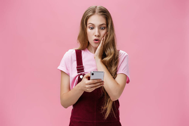 Дівчина дізналася шокуючу правду після того, як прочитала повідомлення в смартфоні, натиснувши руку, щоб щоки в несподіваному вигляді витріщилися стурбованими і безмовними на екрані мобільного телефону, реагуючи на несподівані новини над рожевою стіною
 - Фото, зображення