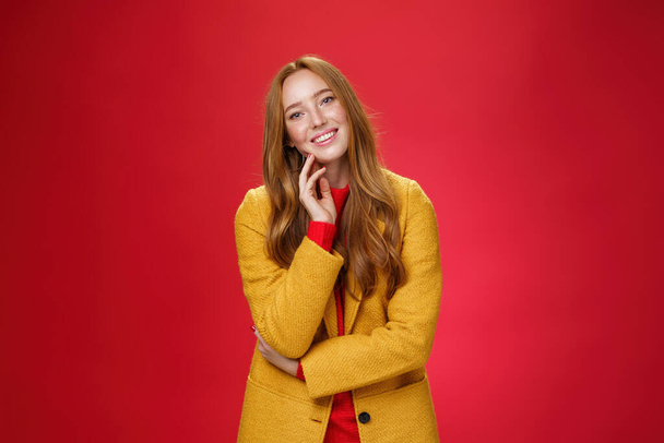 Przystojna, beztroska i szczęśliwa, zrelaksowana ruda dziewczyna w żółtym, stylowym płaszczu dotykająca twarzy i przechylonej głowy, uśmiechnięta pozytywnymi emocjami, pozująca wesoło na czerwonym tle - Zdjęcie, obraz