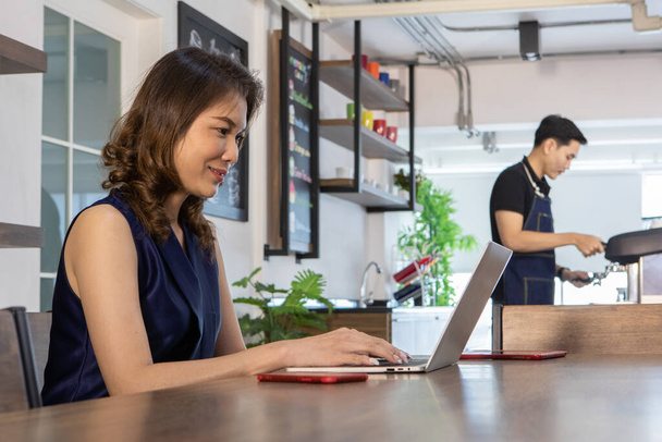Całkiem ładny Azji kobieta freelancer w ciemnym zbiorniku top siedzi w pobliżu licznika barista w kawiarni po południu, spojrzeć na aparat wpisując na laptopie jako szczęśliwy do pracy online biznesu - Zdjęcie, obraz