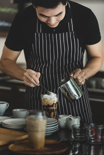 Joven barista asiático en delantal blanco y negro con cuchara para cubrir deliberadamente el café con espuma de leche para la suavidad y la suavidad en el mostrador que rodea con platos, frascos, vasos - Foto, Imagen