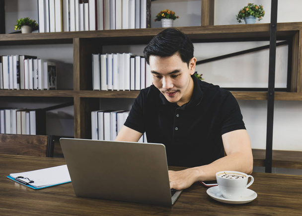 Bel homme asiatique en chemise noire décontractée assis à l'aise sur une table brune avec une tasse de café du matin dans la salle de lecture et profiter des affaires en ligne de travail sur ordinateur portable avec des papiers de note - Photo, image