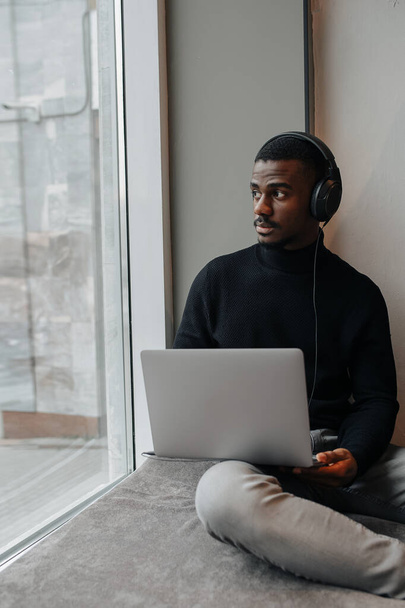 Relaxed black man wearing headphones chilling on a windowsill with laptop, looking from the first floor outside at adjacent street. - Фото, изображение