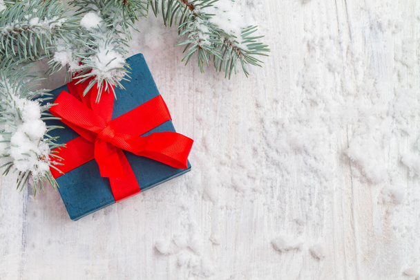 Christmas gift with a blue box with a red ribbon with branches of a green Christmas tree on a white background with snow. - Фото, изображение