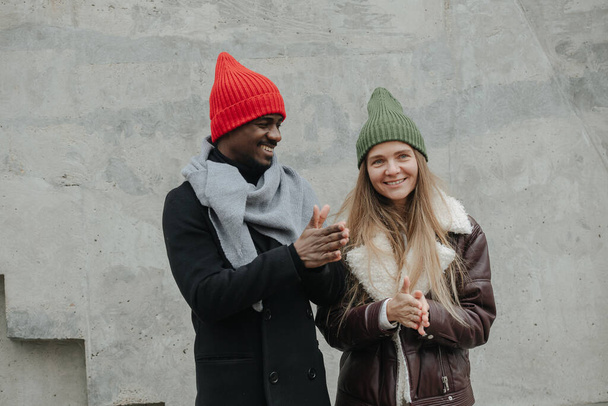 Good interracial opposite sex friends hanging out on the street, rubbing hands for warmth. Both dressed warmly for late autumn. Wearing same fashion knitted hats. - Zdjęcie, obraz