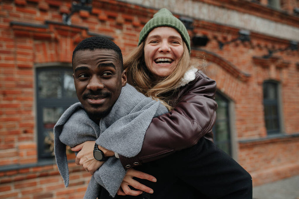 Happy woman riding on her black male friend back on the street. Both dressed warmly for late autumn. Both having a great time, smiling - Foto, Imagem
