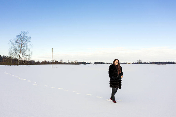 a cheerful woman in a warm fur coat walks across the open field leaving footprint in the snow - Φωτογραφία, εικόνα