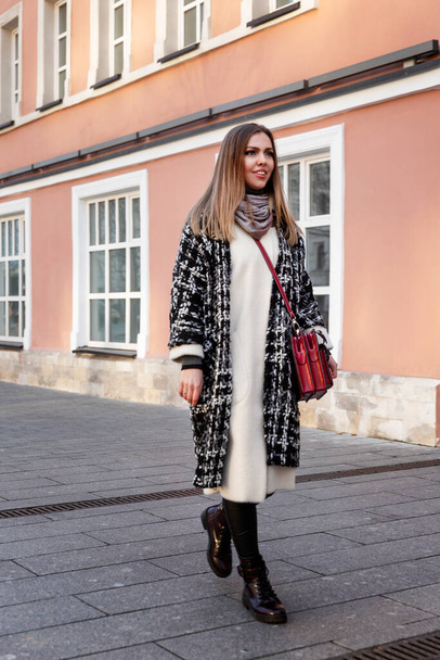 Young woman on the street of the city. Beautiful smiling blonde in a warm stylish coat against the background of a pink house. Full height. Vertical. - Fotografie, Obrázek