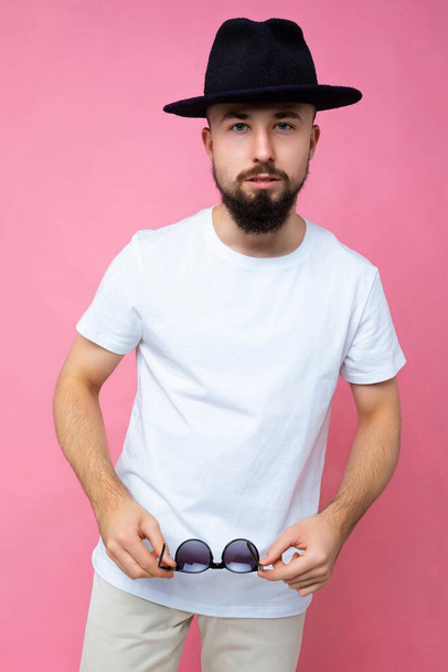 Vertical photo shot of confident brutal good looking young brunet bearded man wearing casual white t-shirt for mockup and stylish black hat poising isolated on pink background with empty space for - Foto, afbeelding