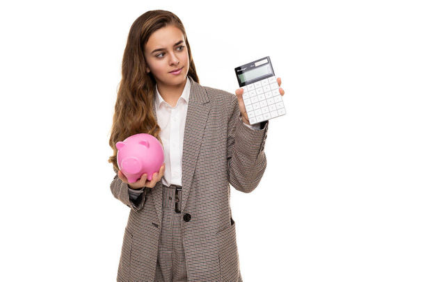 Photo of beautiful cute pretty asking thoughtful brunette teenage girl with long hair in stylish plaid jacket, plaid pants and white shirt holds calculator and pink pig piggy bank isolated on white - Photo, Image