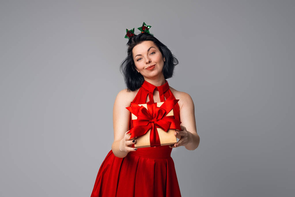 Счастливая юная брюнетка в красном платье позирует на сером фоне с подарочной коробкой, украшенной красной лентой. Праздники, праздники и образ жизни - Фото, изображение