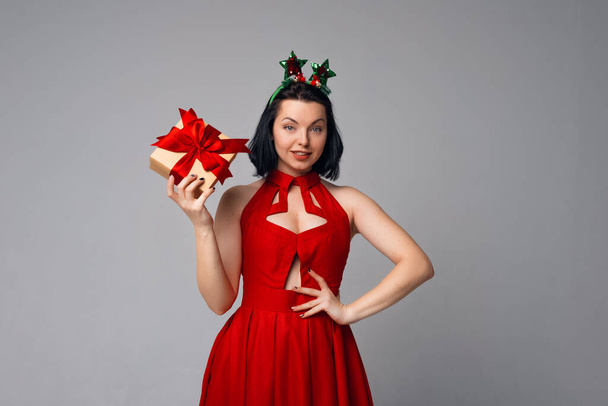 Curious young brunette woman in red dress posing on gray background with gift box decorated with red ribbon. Holidays, celebration and lifestyle concept - Foto, Bild