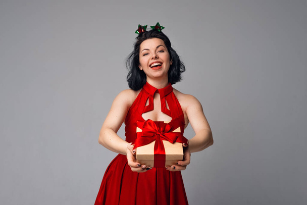 Όμορφη χαμογελαστή γυναίκα σε κόκκινο φόρεμα κρατώντας κουτί δώρου διακοσμημένα με κόκκινη κορδέλα. Έννοια διακοπών, γιορτής και τρόπου ζωής - Φωτογραφία, εικόνα