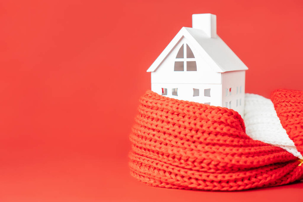 Fehér játékház piros sálba csomagolva, piros háttérrel. Fűtés vagy szigetelés épületek vagy házak koncepciója. Fénymásolási hely - Fotó, kép