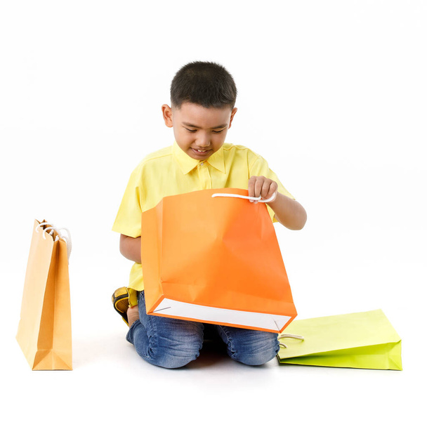 Cutout portrait of little Asian boy on yellow shirt and blue jeans happily sit beside attractive shopping paper bag and excitedly open orange one to find interesting sale or surprised product inside - 写真・画像
