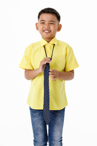 Ausgeschnittenes Porträt eines smarten kleinen asiatischen Jungen auf gelbem Hemd, blauer Jeans, lächelnd stehend und genüsslich Krawatte als formelle Uniform tragend, die sich auf eine glückliche Arbeit als junger Geschäftsmann oder Büroangestellter vorbereitet. - Foto, Bild