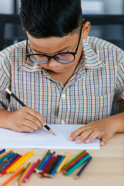 Smart young Asian boy wearing glasses and grey checked shirt sitting on studying desk of tablet, mobile phone, and colorful pencils in reading room and concentrate on drawing on white paper - Photo, Image
