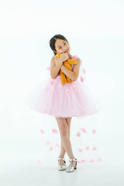 Portrét izolované studio záběr malé asijské roztomilé balerína dítě v růžové krásné elegance růže květinový balet šaty stojící objímání oranžové nadýchaný medvídek panenka v náručí v přední bílé pozadí. - Fotografie, Obrázek