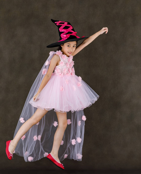 Portrait-Studioaufnahme von kleinen süßen asiatischen Kind in rosa Hexenkleid Kostüm mit schwarzem Hut Blick in die Kamera posiert fliegende Geste mit Spaß auf Halloween traditionelles Fest. - Foto, Bild