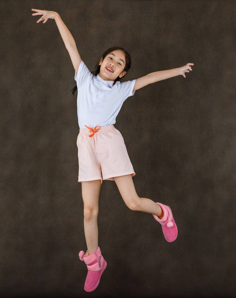 Portrét studio záběr na malé roztomilé hravé šťastný asijské dívka nosí bílou košili a růžové šortky a boty skákání vysoko ukazující mírové znamení se dvěma prsty nad hlavou v přední části hnědé stěny pozadí. - Fotografie, Obrázek