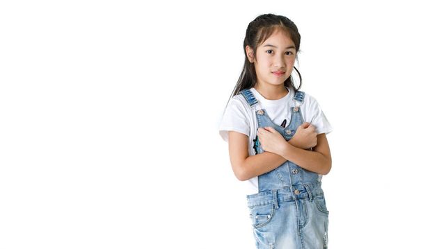 Портрет крупным планом изолированная студия снимок азиатской маленькой милой девочки носит футболку и крутые джинсы джинсовой юбки комбинезон стоя улыбаясь смотреть на камеру скрещенные руки на груди перед белым фоном. - Фото, изображение