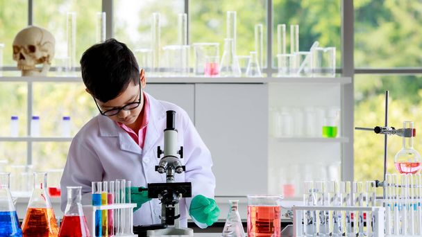 Asiático colegial en gafas y bata de laboratorio examinando sustancia química a través del microscopio mientras conduce experimento científico en laboratorio escolar. - Foto, Imagen