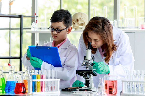 Okul öğretmeni mikroskopla kimyasal örnekleri incelerken okul çocuğu da laboratuarda kimya araştırması sırasında sonuçları not alıyor. - Fotoğraf, Görsel