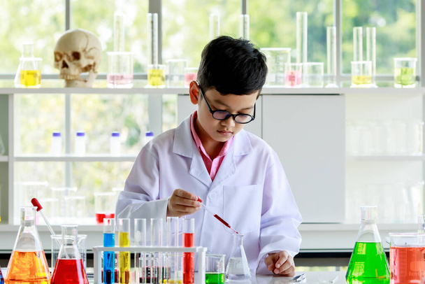 Ernstige aziatische jongen in lab jas en bril met druppelaar en buis mengen kleurrijke reagentia tijdens het uitvoeren van wetenschappelijk onderzoek in moderne school laboratorium. - Foto, afbeelding