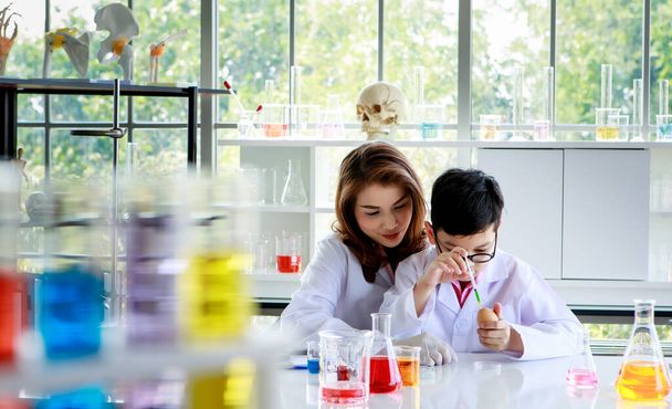 Ασιάτης σχολιαρόπαιδο ανάμειξη πολύχρωμα υγρά κατά τη διεξαγωγή πειράματος με τη βοήθεια του καθηγητή κατά τη διάρκεια μαθήματος χημείας στο εργαστήριο - Φωτογραφία, εικόνα