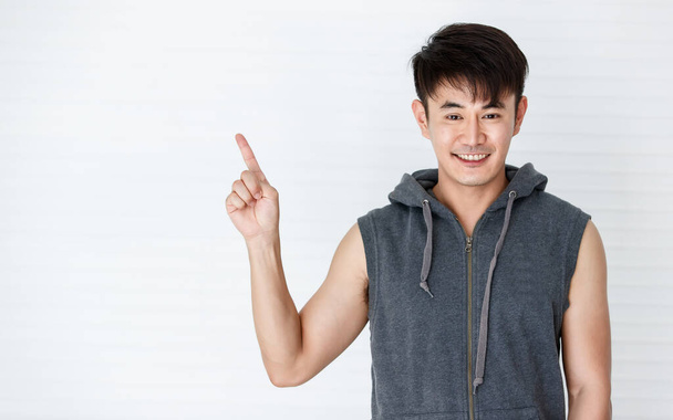 asiático guapo ajuste sonriente hombre sosteniendo punto el dedo para copiar espacio usando sin mangas camiseta gris sportswear en blanco fondo - Foto, imagen